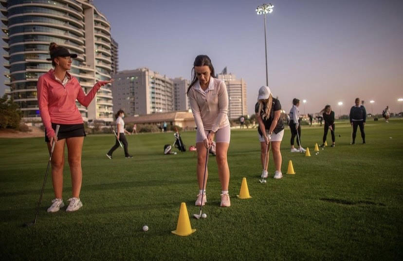 A Chicks With Sticks coach guiding women golfers through a chipping drill at dusk in Dubai